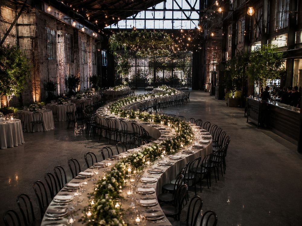 Modern warehouse wedding reception featuring curved tables, greenery and romantic lighting.