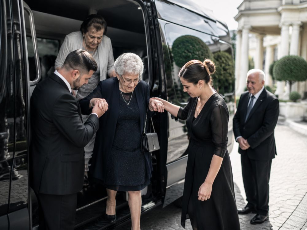 Elderly wedding guests receiving assistance from a luxury transport vehicle near the venue entrance.